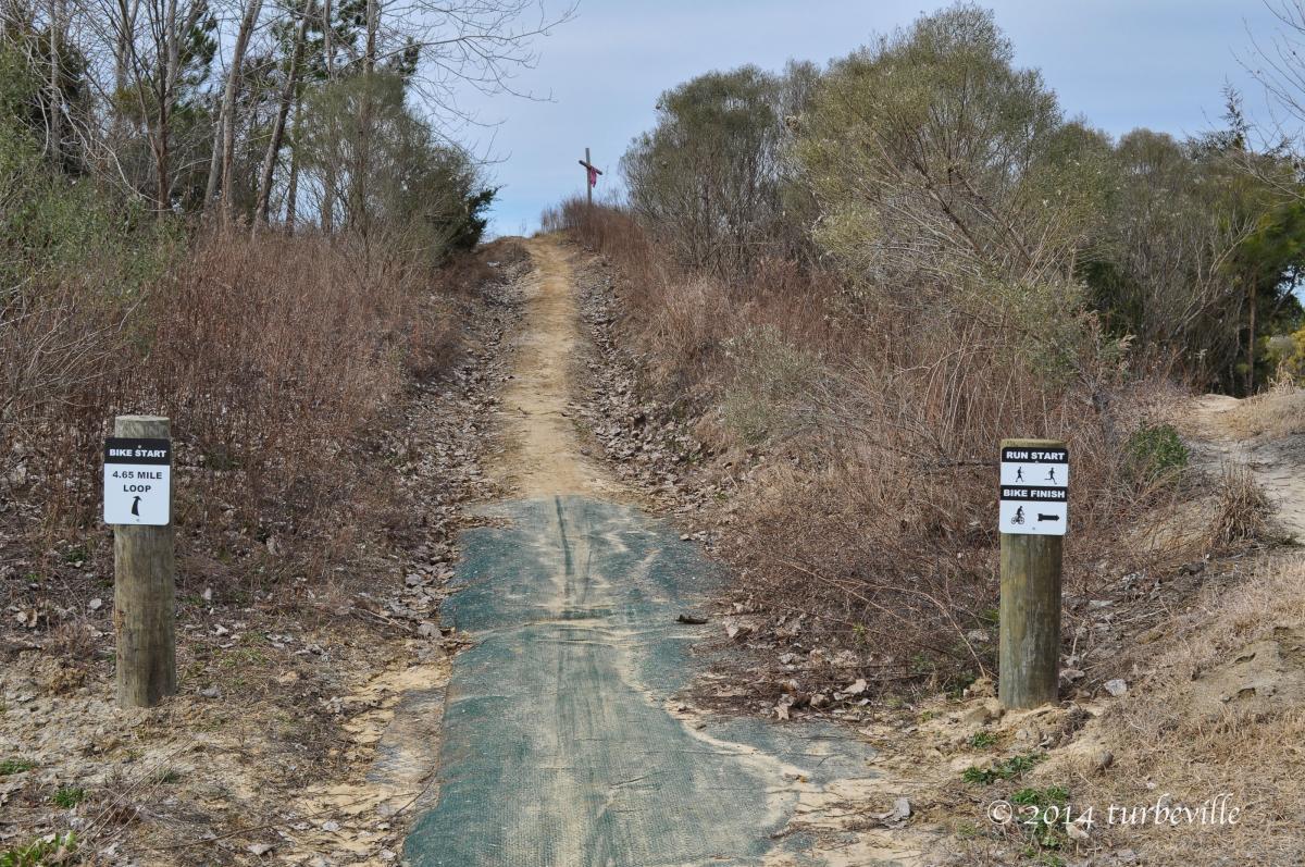 A dirt path leading into a forested area with signs marking the start of a 4.65-mile bike loop and a run start. The path is lined with sparse vegetation and trees, and the sky is overcast. Horry County Bike Run Park mountain bike trail.