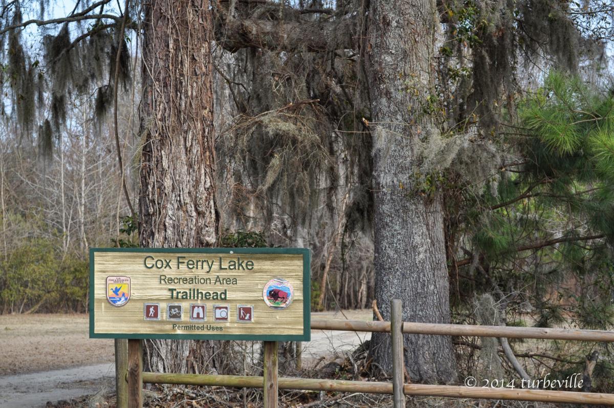 Signage for Cox Ferry Lake Recreation Area Trailhead, featuring permitted use icons for hiking, biking, and walking, surrounded by trees and natural foliage. Jackson Bluff Trail System mountain bike trail.