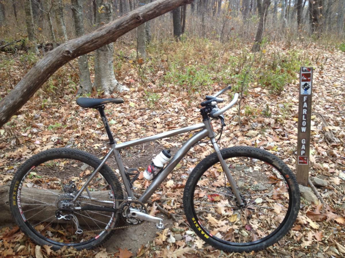 A silver mountain bike parked on a trail surrounded by fallen leaves and trees. A wooden log leans across the trail, and a sign labeled "FARLOW GAP" is positioned nearby. The scene depicts an autumn setting with varying shades of brown and green foliage. Farlow Gap mountain bike trail.
