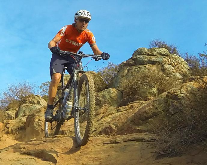 A mountain biker in an orange jersey and helmet navigates a rocky terrain with determination, surrounded by scrub vegetation under a clear blue sky. Dust is kicked up as the biker rides over uneven ground, showcasing skill and adventure in outdoor cycling. Rancho La Costa mountain bike trail.
