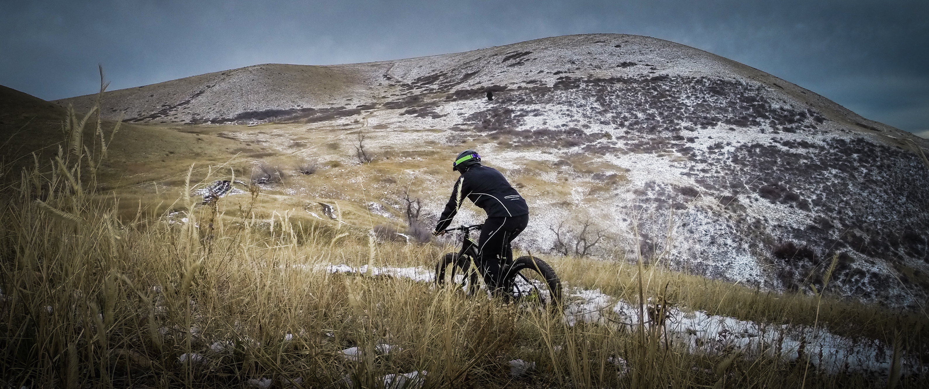 A cyclist riding a fat bike on a snowy, grassy hill with rolling terrain in the background under a cloudy sky.