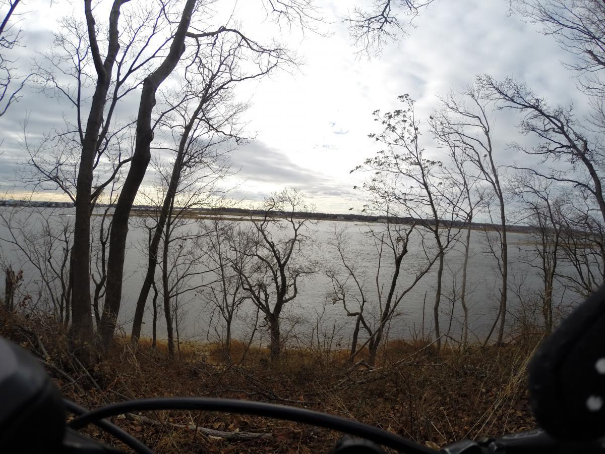 A scenic view of a river framed by bare winter trees, with a cloudy sky above. The image shows a sense of tranquility and nature, as seen from a vantage point that includes parts of a bicycle in the foreground. Hartshorne Woods Park mountain bike trail.