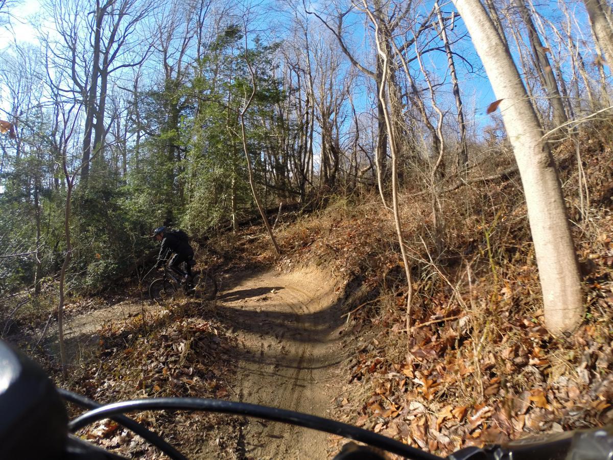 A mountain biker navigating a curved dirt trail through a wooded area, with bare trees and green shrubs in the background. The scene is set on a sunny day, highlighting the natural landscape. Hartshorne Woods Park mountain bike trail.