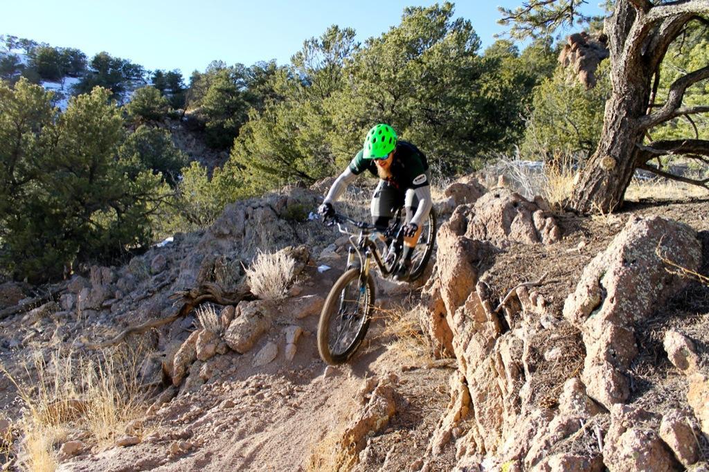 A mountain biker navigating a rocky trail surrounded by trees and brush, wearing a bright green helmet and a black cycling outfit. The terrain is uneven and challenging, showcasing the rugged beauty of the natural landscape. Sand Dunes mountain bike trail.
