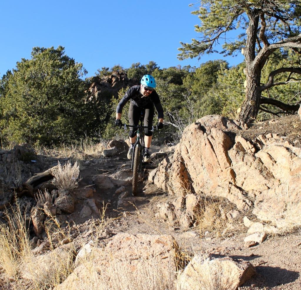 A mountain biker navigating a rocky trail during a sunny day, surrounded by trees and shrubs. Sand Dunes mountain bike trail.