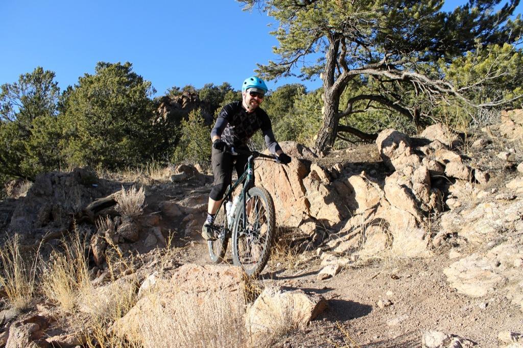 A cyclist in a blue helmet rides a mountain bike along a rocky trail in a forested area, surrounded by tall trees and dry grass under a clear blue sky. Sand Dunes mountain bike trail.