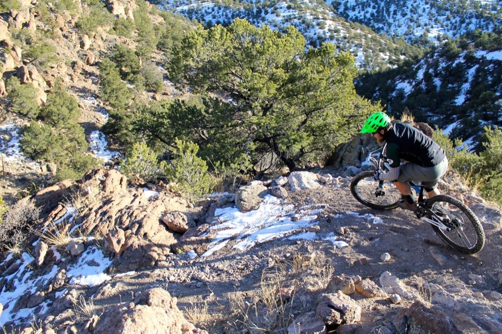 A mountain biker navigating a rocky trail in a mountainous landscape with greenery and patches of snow, wearing a bright green helmet and athletic gear. Sand Dunes mountain bike trail.