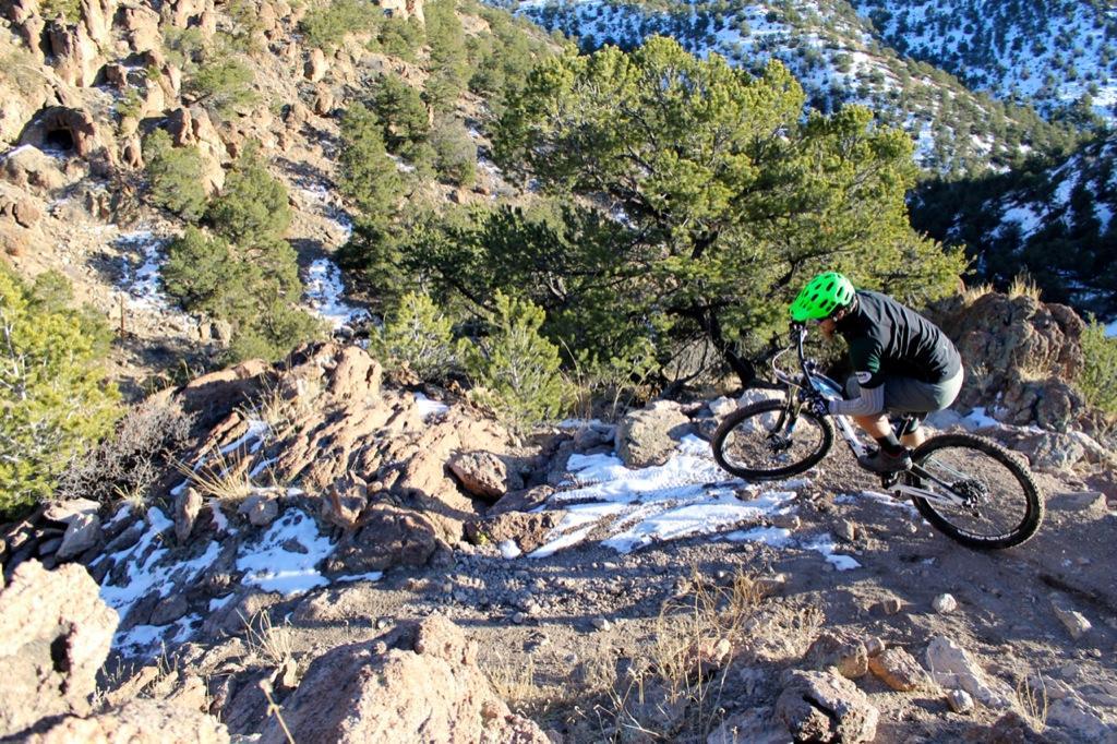 A mountain biker wearing a green helmet navigates a rocky trail surrounded by trees and patches of snow in a mountainous landscape. The rider is in motion, showcasing a dynamic moment as they maneuver down the incline. Sand Dunes mountain bike trail.