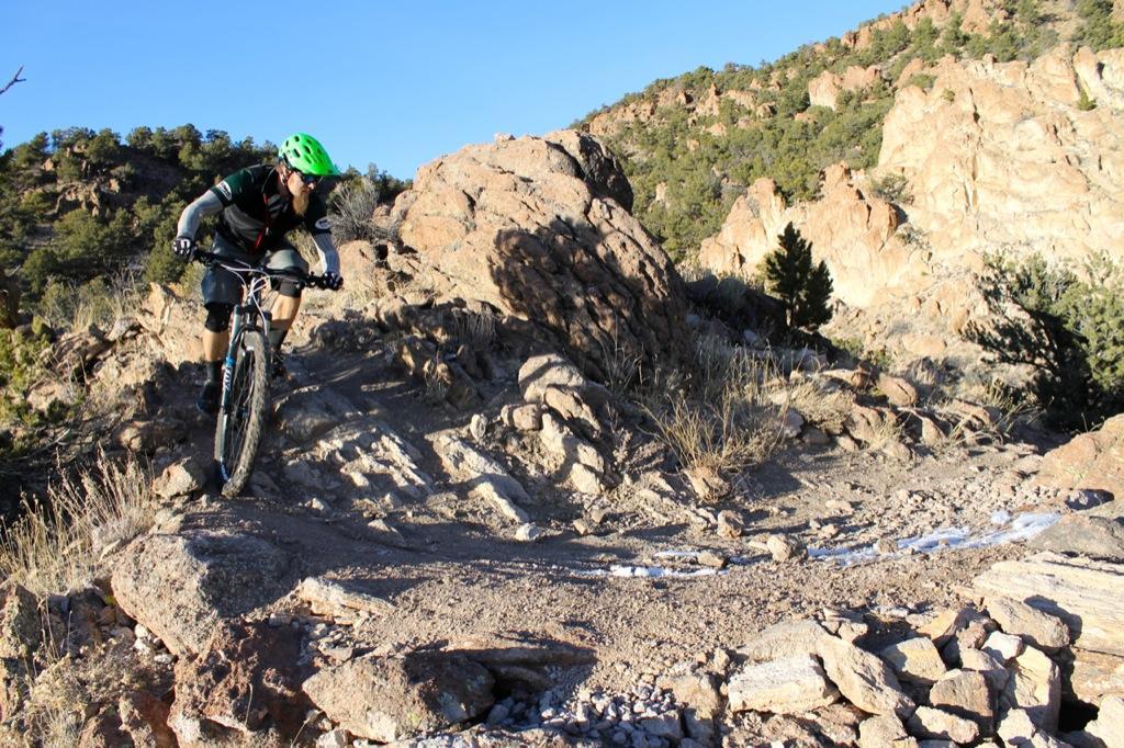 A mountain biker navigates a rocky trail in a rugged outdoor landscape, surrounded by hills and trees under a clear blue sky. Sand Dunes mountain bike trail.