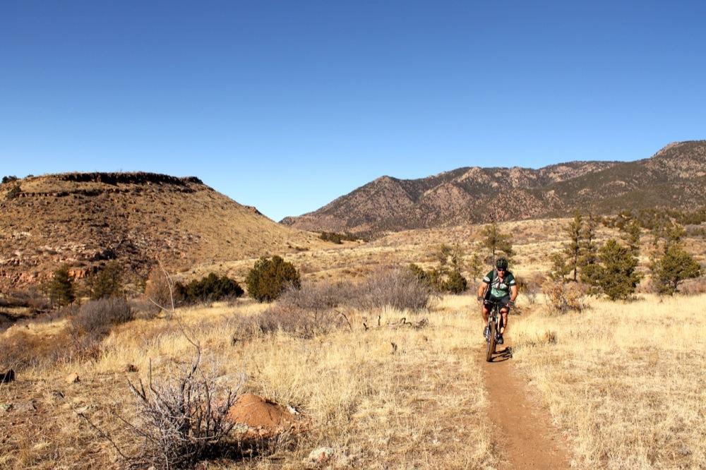 A person riding a mountain bike on a dirt trail through a grassy landscape with low hills and distant mountains under a clear blue sky. Oil Well Flats mountain bike trail.