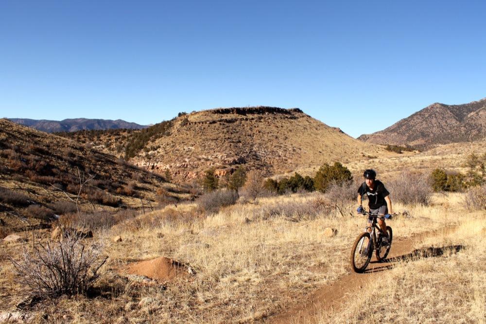 A person riding a mountain bike along a dirt trail in a hilly landscape, surrounded by dry grass and sparse vegetation under a clear blue sky. Oil Well Flats mountain bike trail.