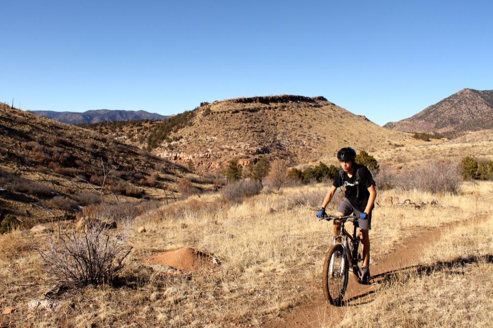 A person riding a mountain bike on a dirt trail through a rocky, arid landscape with rolling hills and sparse vegetation under a clear blue sky. Oil Well Flats mountain bike trail.