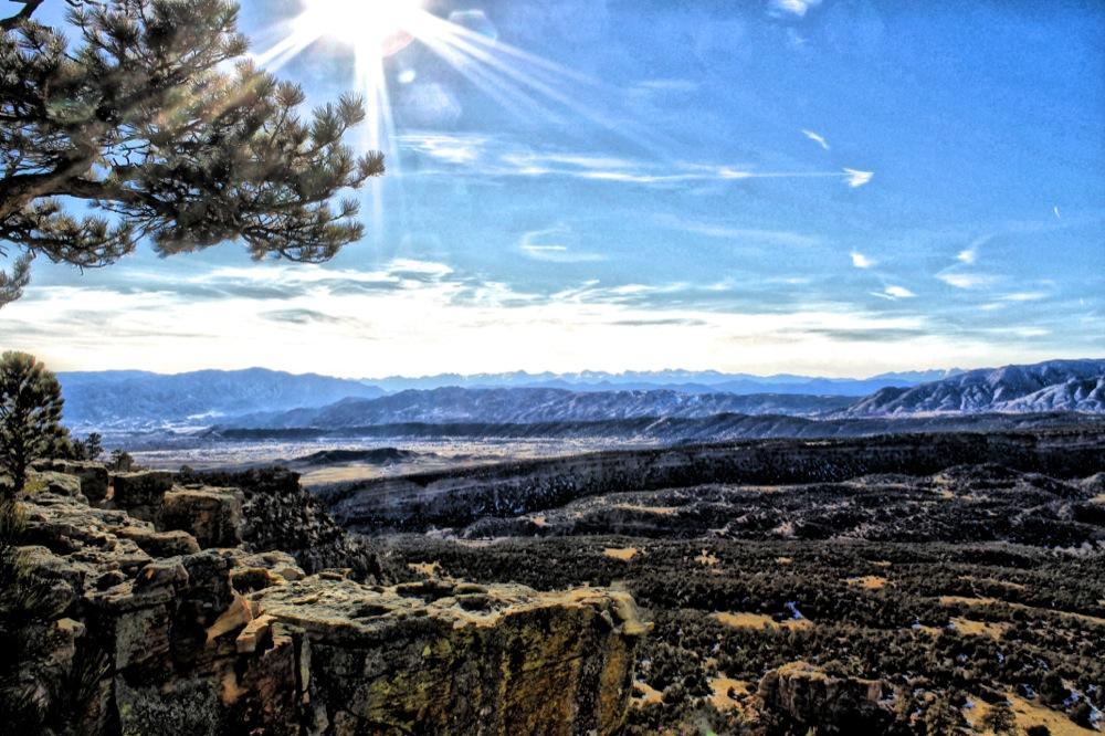 A scenic view from a rocky ledge overlooking a vast valley and distant mountains beneath a bright blue sky with scattered clouds. The sunlight shines brightly, illuminating a pine tree in the foreground. The landscape features a mix of greenery and rocky terrain, showcasing the beauty of nature. Oil Well Flats mountain bike trail.