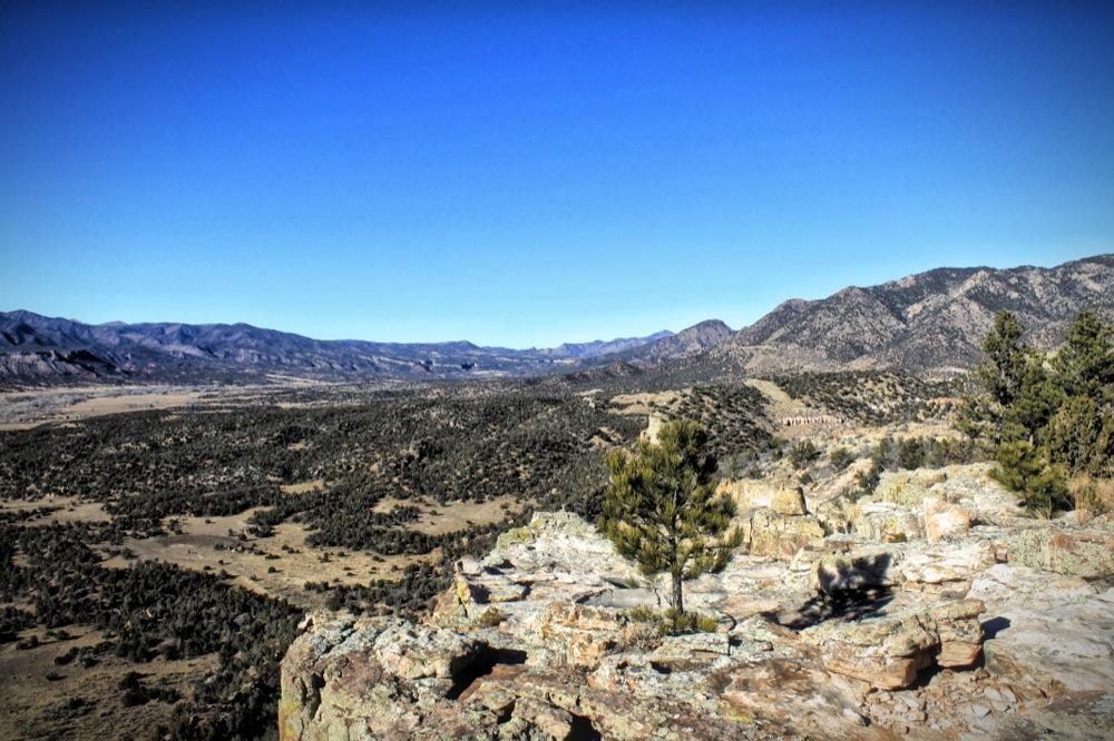 A panoramic view of a mountainous landscape under a clear blue sky, featuring rocky outcrops and scattered evergreen trees. The foreground shows rocky terrain leading to a vast expanse of hills and valleys in the distance, highlighting the natural beauty of the scenery. Oil Well Flats mountain bike trail.