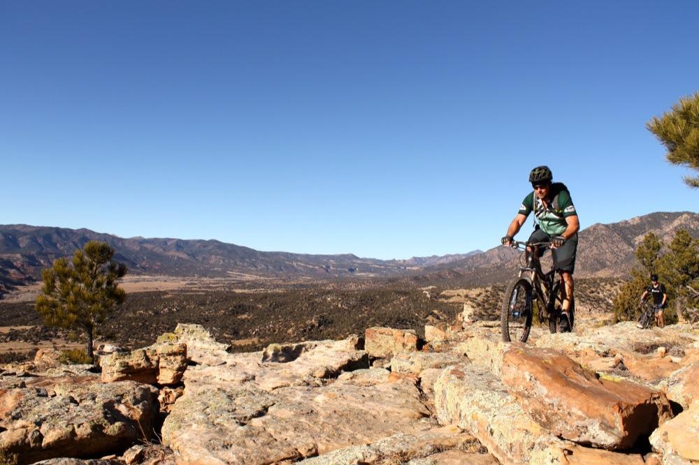 Mountain biker navigating rocky terrain with a scenic landscape of mountains and valleys in the background under a clear blue sky. Oil Well Flats mountain bike trail.