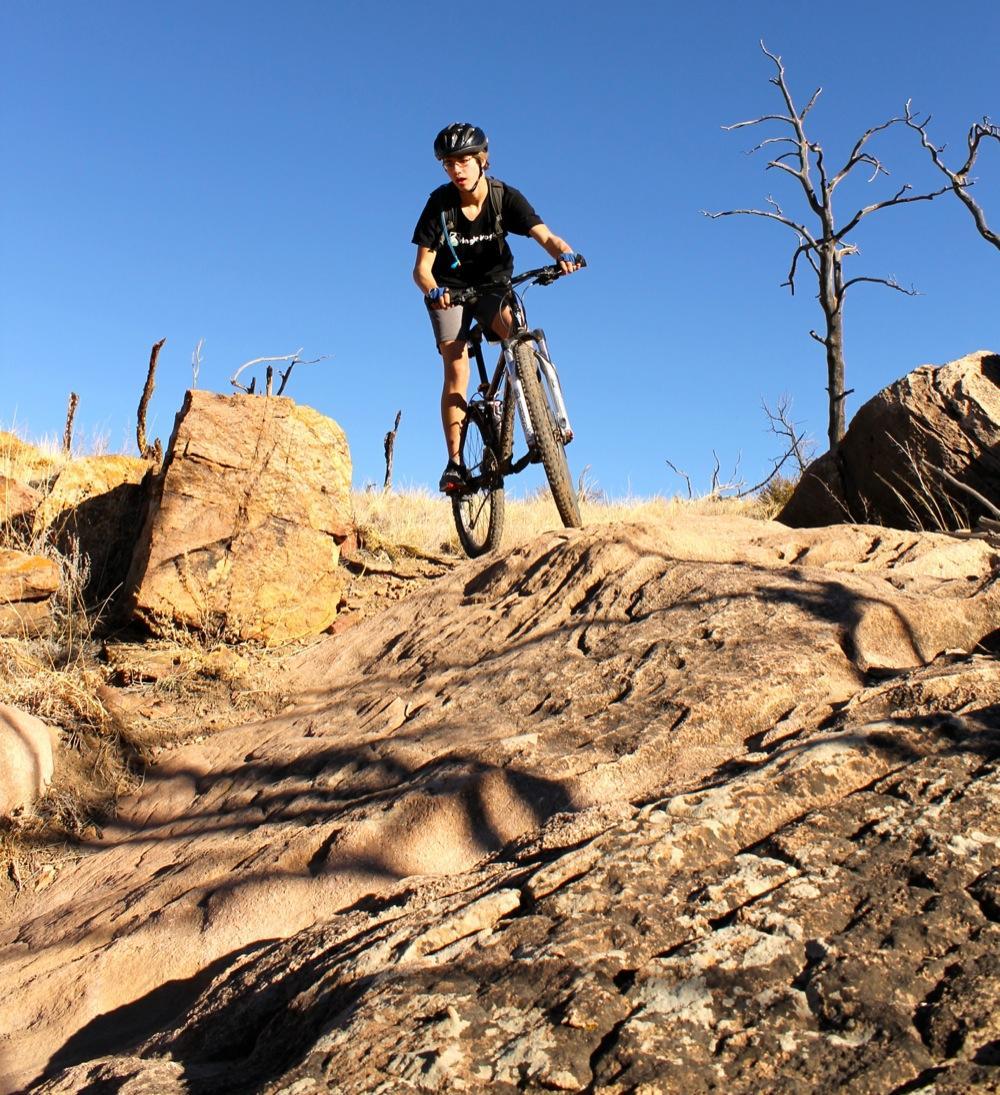 A person riding a mountain bike over rocky terrain against a clear blue sky. The cyclist is wearing a helmet and a t-shirt, showcasing an adventurous spirit while navigating the uneven landscape. Dry grass and sparse trees are present in the background, emphasizing an outdoor setting. Oil Well Flats mountain bike trail.