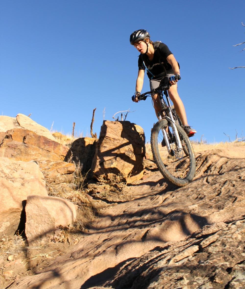 A person riding a mountain bike down a rocky slope, with a clear blue sky in the background. The rider is wearing a black helmet and a black shirt, focusing intently as they navigate the rough terrain. Large boulders and dry grass are visible around them. Oil Well Flats mountain bike trail.
