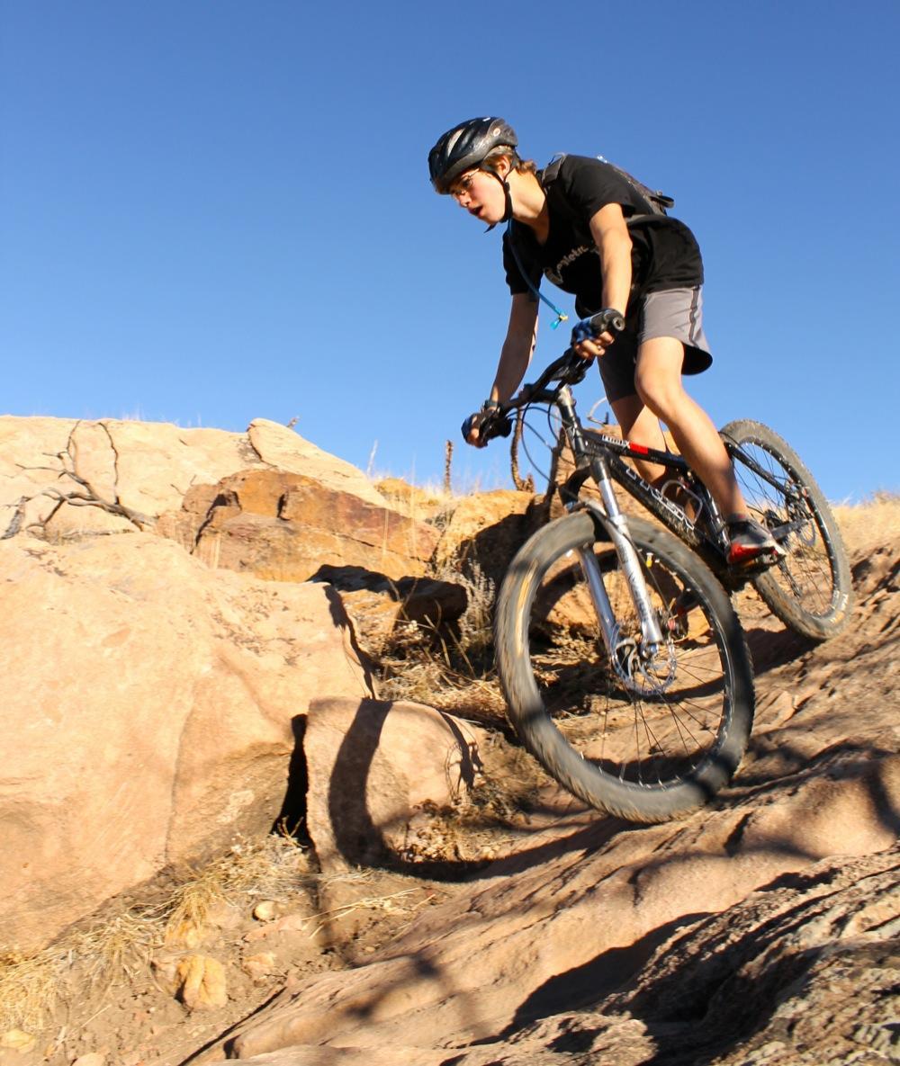 A young person riding a mountain bike down a rocky trail under a clear blue sky. The cyclist is focused and leaning forward, navigating the uneven terrain with determination. The landscape features natural rock formations and dry grass. Oil Well Flats mountain bike trail.
