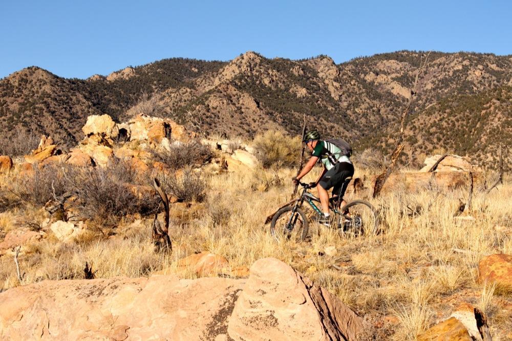 A mountain biker riding on a rocky trail in a rugged landscape, surrounded by dry grass and scattered boulders, with mountains in the background under a clear blue sky. Oil Well Flats mountain bike trail.
