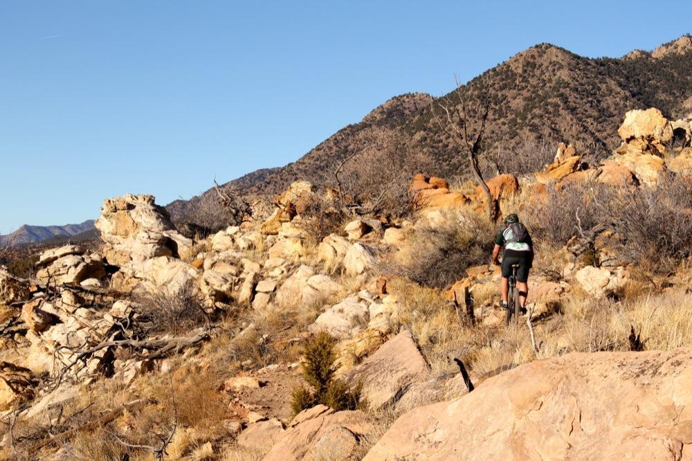 A mountain biker navigates a rocky, arid trail surrounded by boulders and dry vegetation, with mountainous terrain in the background under a clear blue sky. Oil Well Flats mountain bike trail.