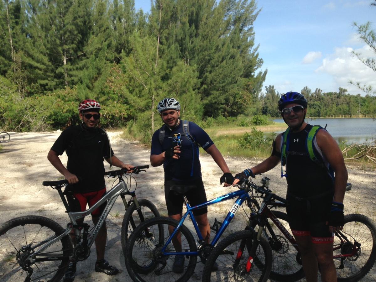 Three mountain bikers pose for a photo on a dirt path surrounded by tall green trees. They each wear helmets and cycling gear, with two holding their bikes while the third holds a water bottle. A calm body of water can be seen in the background under a clear blue sky. Markham Park mountain bike trail.