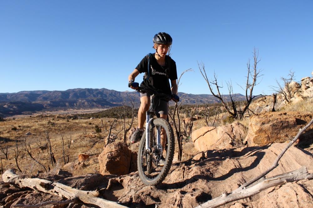 A young person riding a mountain bike over rocky terrain, surrounded by a dry, mountainous landscape under a clear blue sky. Oil Well Flats mountain bike trail.