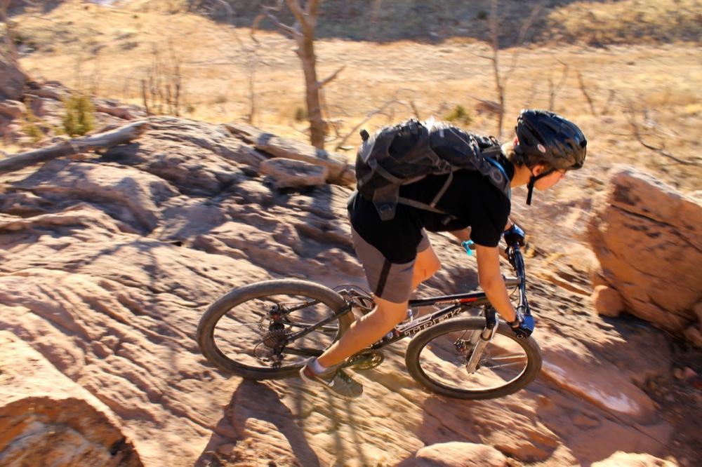 A person riding a mountain bike on rocky terrain, leaning forward while navigating the uneven surface. The background features dry landscape with sparse vegetation and rocky formations under a clear sky. Oil Well Flats mountain bike trail.