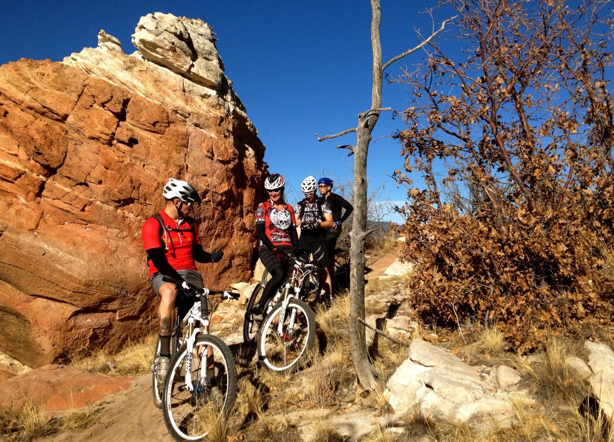 Four mountain bikers, wearing helmets and cycling gear, pause on a rocky trail surrounded by red rock formations and sparse vegetation. The clear blue sky provides a vibrant backdrop as they share a moment in the sunny outdoor setting. Oil Well Flats mountain bike trail.