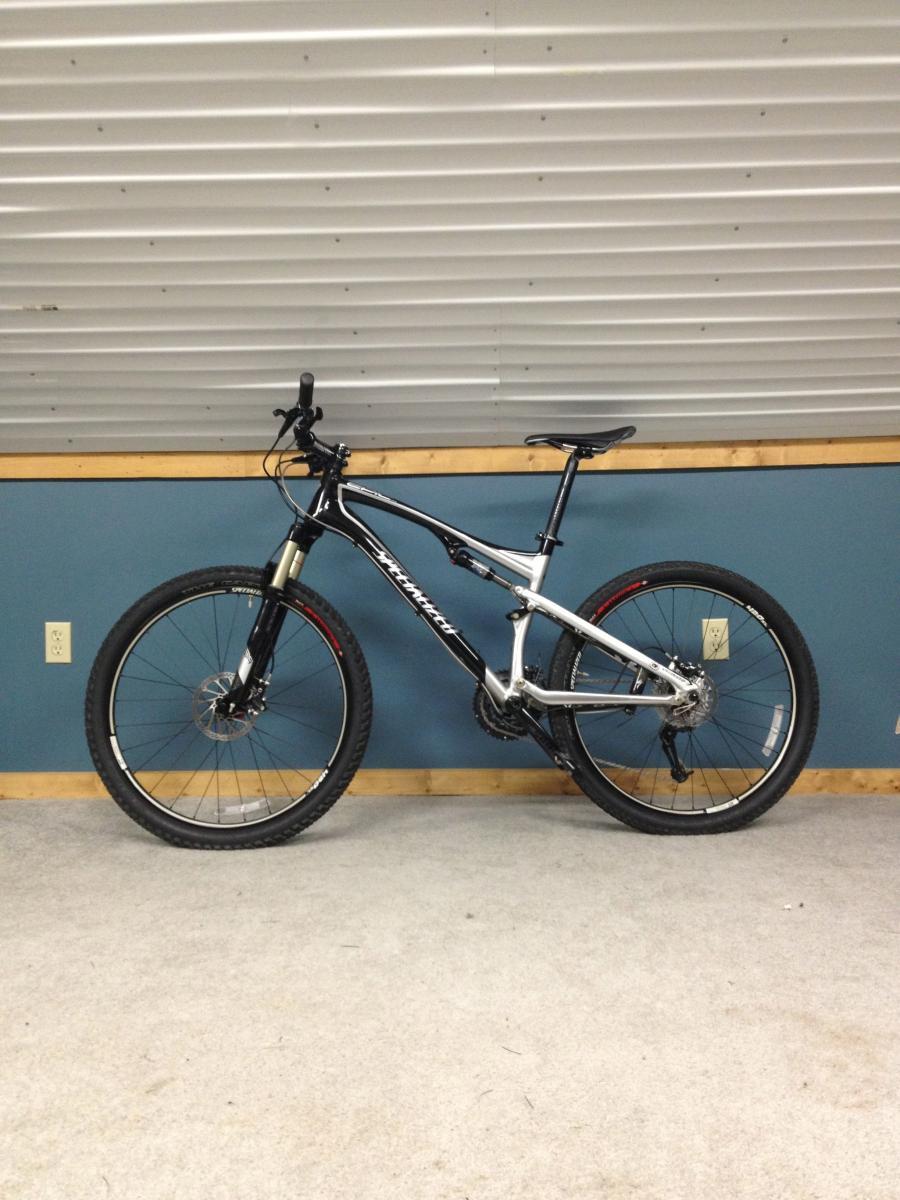 Specialized Epic Comp: Mountain bike standing upright on a carpeted floor against a blue wall with a metal panel background. The bike features a black and silver frame with thick tires suitable for off-road riding.