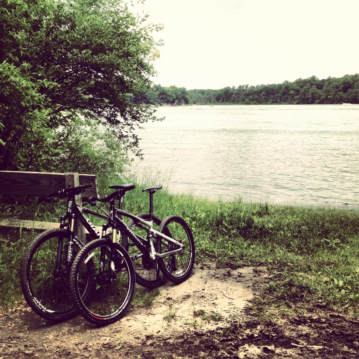 Two bicycles are parked near a riverbank, surrounded by grass and greenery. The calm water of the river reflects the lush landscape, while a wooden fence provides a clear view of the scenery. The atmosphere is serene and inviting, suggesting an ideal spot for outdoor activities. Potawatomi trail mountain bike trail.