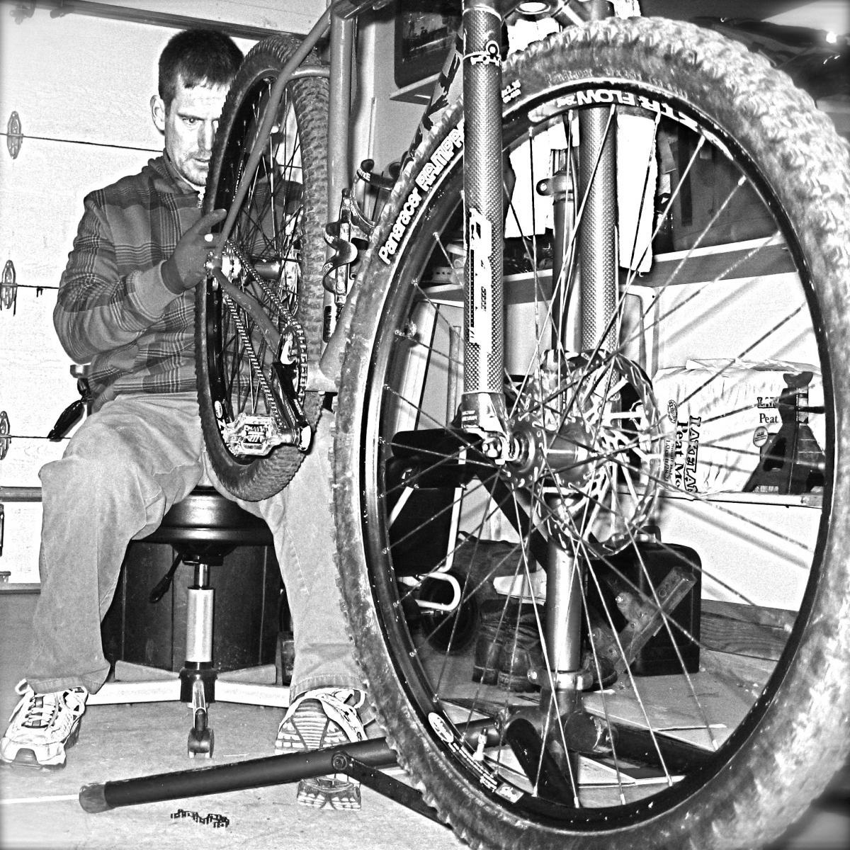 A person is sitting on a stool in a garage, working on a mountain bike. The bike's rear wheel is elevated on a stand, and the individual is focused on adjusting the wheel. The image is in black and white, with various bike parts and tools visible in the background.