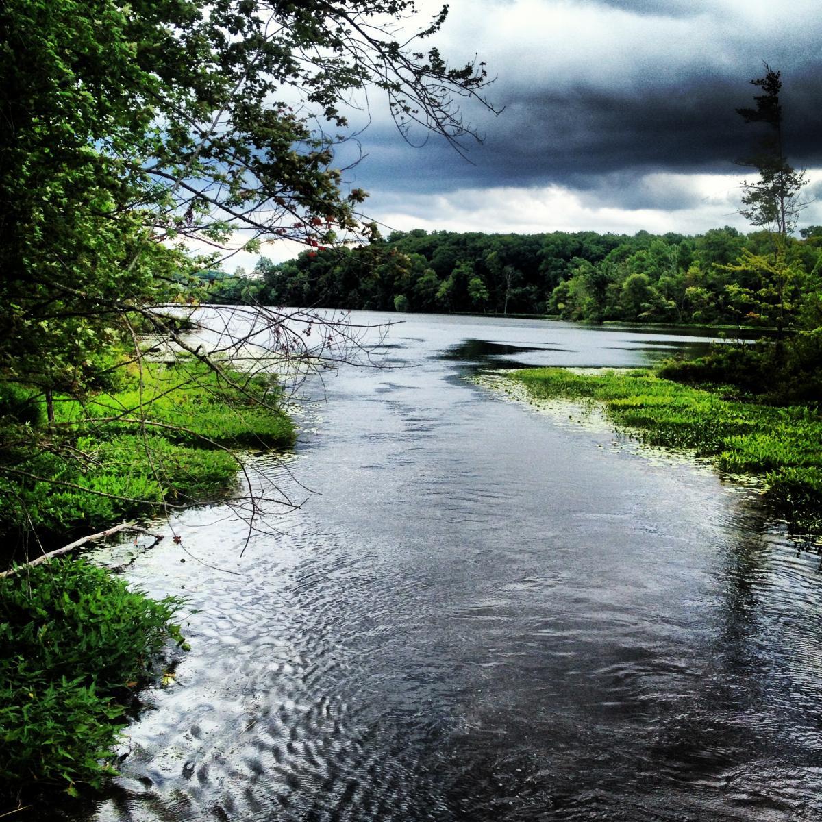 A tranquil river scene surrounded by lush greenery, with dark storm clouds looming overhead. The water reflects the overcast sky and the surrounding landscape, while patches of vegetation border the riverbanks. Potawatomi trail mountain bike trail.