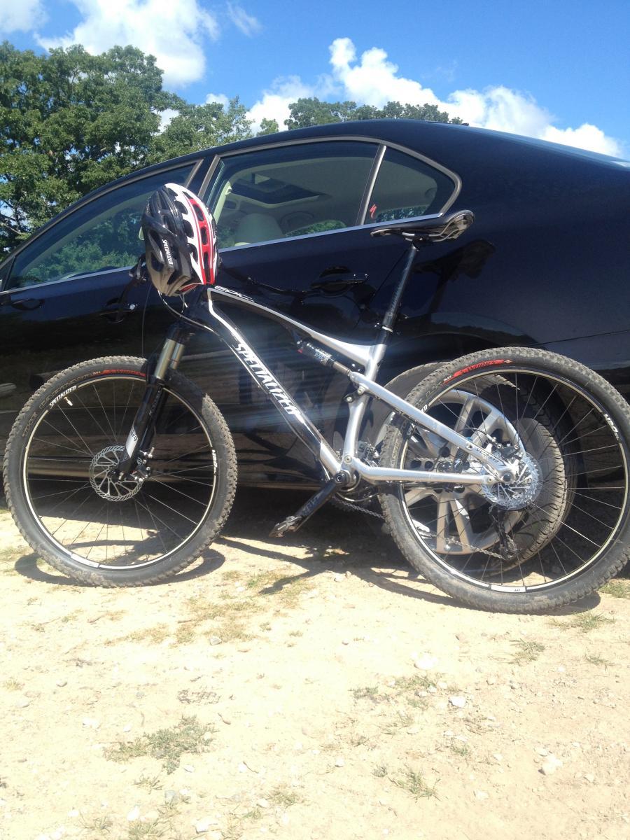 Specialized Epic Comp: A silver mountain bike with a red and black helmet resting on its handlebars, parked on a dirt surface next to a black car. The background features a clear blue sky with a few clouds and green trees.
