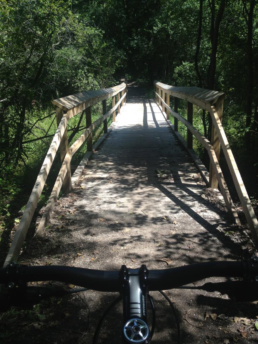 A wooden bridge surrounded by lush greenery, viewed from the perspective of a bicycle handlebar. The path leads into a shaded trail, with dappled sunlight filtering through the trees. The bridge features wooden railings and a textured, gravelly surface. Brighton Rec Area mountain bike trail.