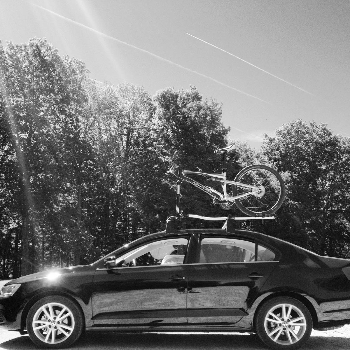 Specialized Epic Comp: A black and white image of a car parked on a sunny day with a bicycle mounted on a roof rack. Trees are visible in the background, and the sky shows a few contrails.