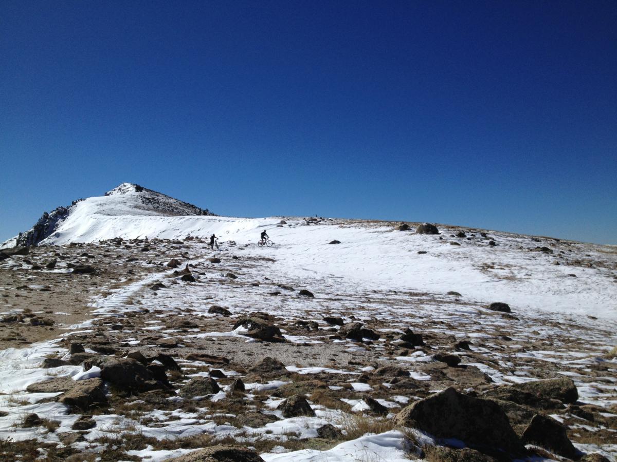 Two cyclists navigating a snowy mountain trail under a clear blue sky. The terrain features rocky patches and a snow-covered path leading towards a peak in the background. Monarch Crest Trail mountain bike trail.