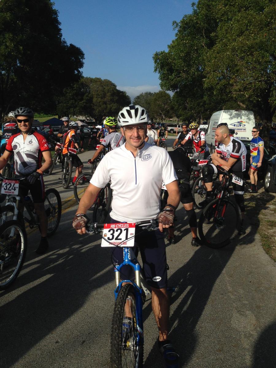 A group of cyclists gathered at an outdoor event, preparing for a race. In the foreground, a smiling man in a white shirt and a helmet stands next to his blue mountain bike, displaying his race number, 321. Other cyclists in various jerseys and helmets are visible in the background, with trees and a clear blue sky overhead. Markham Park mountain bike trail.