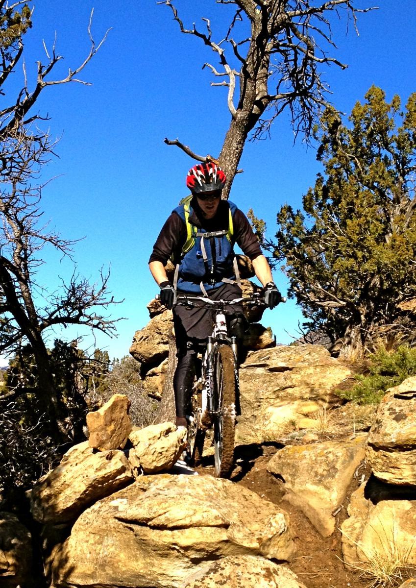 A mountain biker navigates a rocky trail surrounded by trees under a clear blue sky. The rider is wearing a helmet, gloves, and a backpack, crouched over the handlebars as they maneuver over stones. Oil Well Flats mountain bike trail.