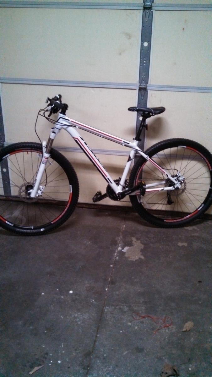 Trek Cobia: A white mountain bike with red accents parked inside a garage, displayed against a beige wall. The bike features thick tires, a curved frame, and a comfortable seat, with tools and debris visible on the garage floor.