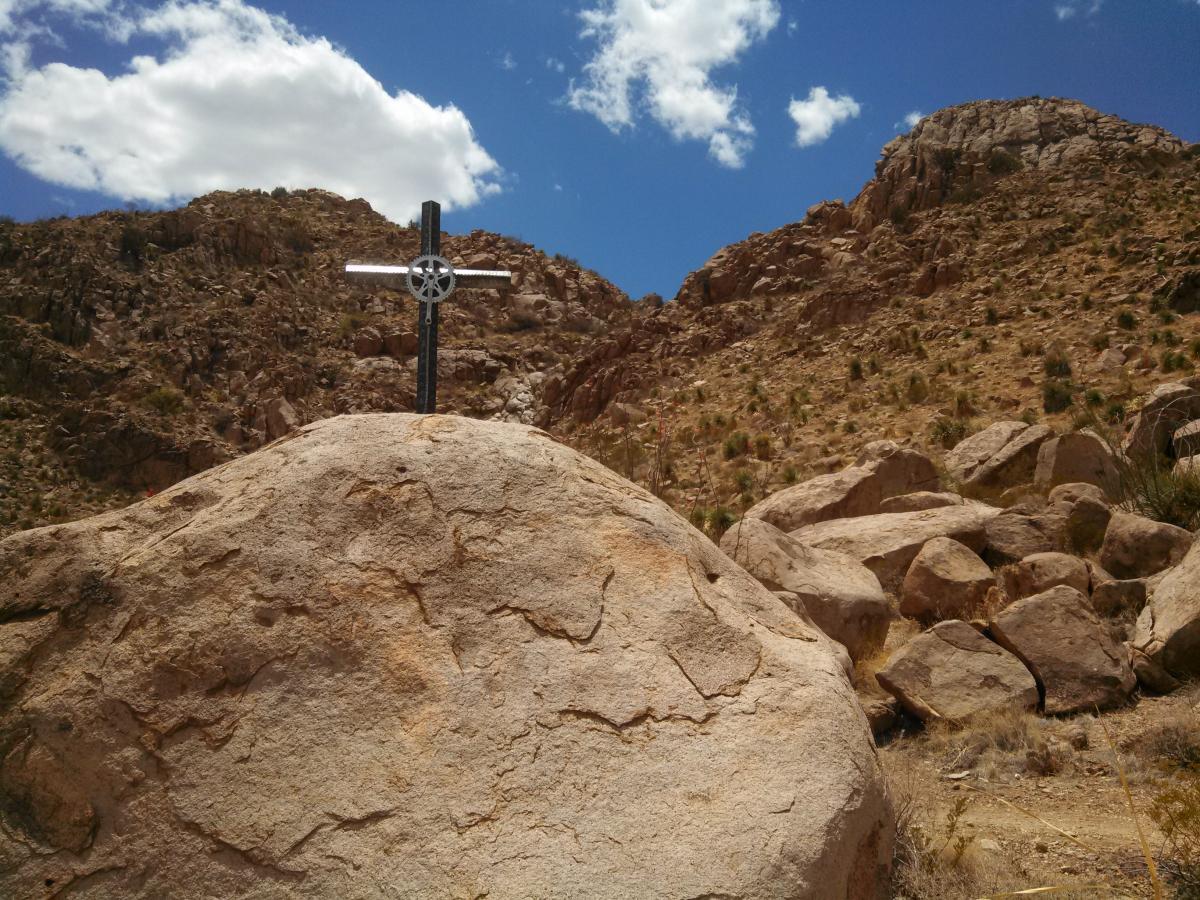 A metal cross on a large rock, set against a backdrop of rugged mountains and a blue sky with white clouds. The rocky terrain is dry and arid, with scattered vegetation in the foreground. Dona Ana mountain bike trail.
