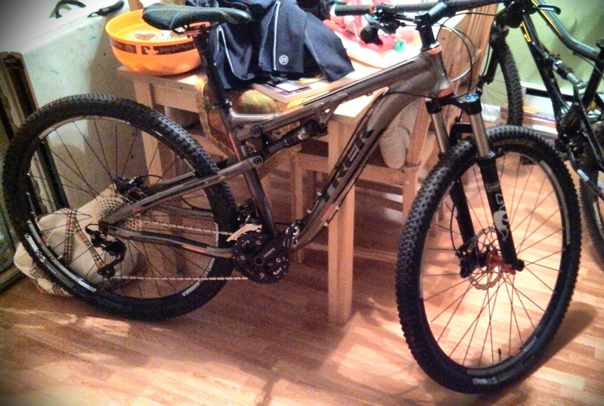 Trek Superfly 100 AL Elite: A mountain bike with a gray frame and thick tires is leaning against a table. The bike features a suspension system, chain, and disc brakes. In the background, there are kitchen items and a woven blanket on a chair. The setting is indoors, with wooden flooring and a casual atmosphere.