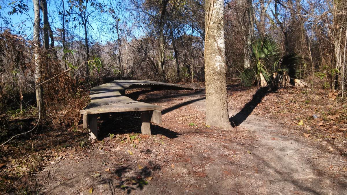 A winding wooden boardwalk surrounded by trees and underbrush, leading to a dirt path through a forest in daylight. Bonnet Carre Spillway Trail mountain bike trail.