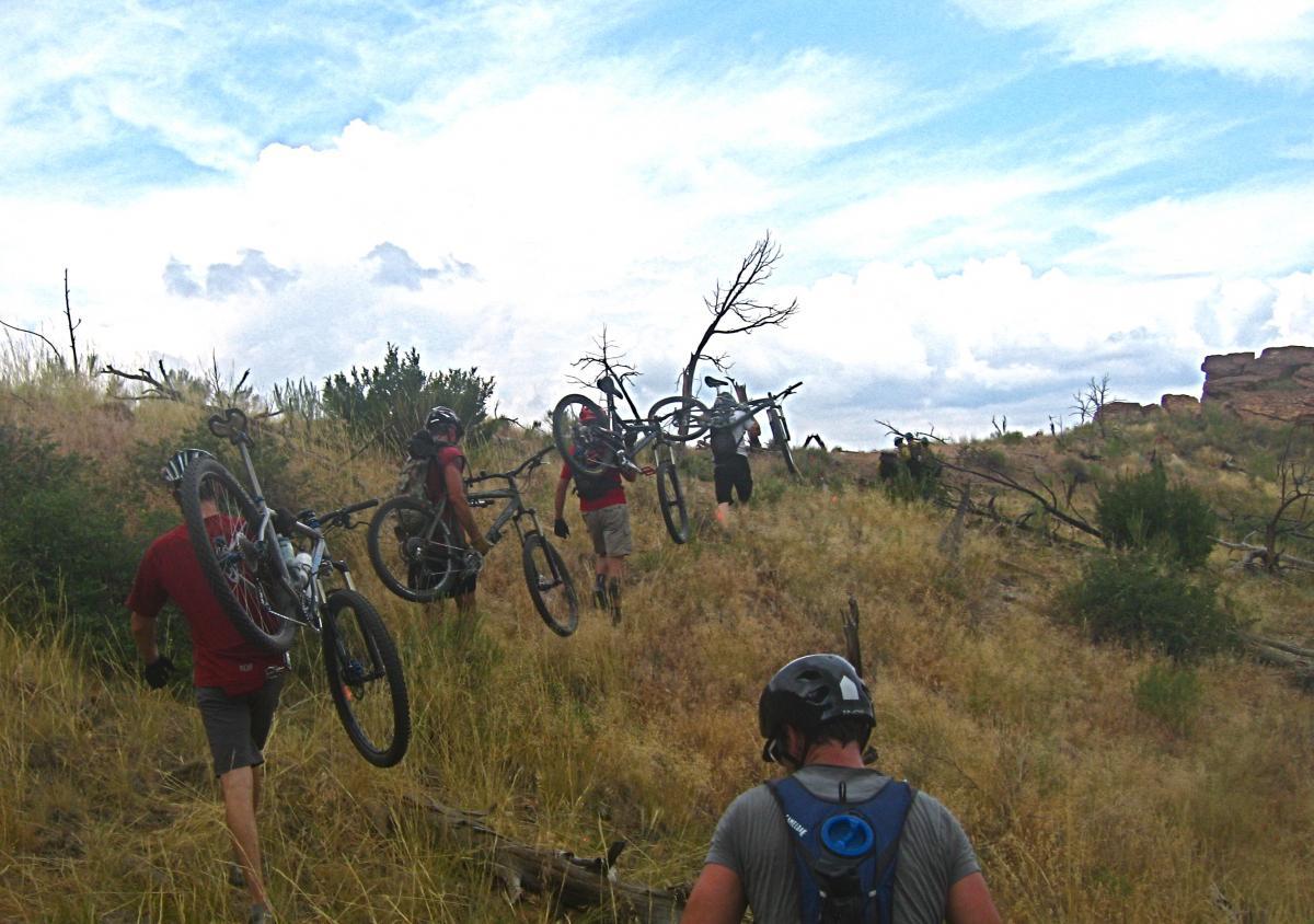 A group of mountain bikers walking uphill on a grassy trail, carrying their bicycles over their shoulders. The landscape features sparse vegetation, scattered trees, and a cloudy sky. The riders are dressed in casual biking attire and helmets, with some visible backpacks. Oil Well Flats mountain bike trail.