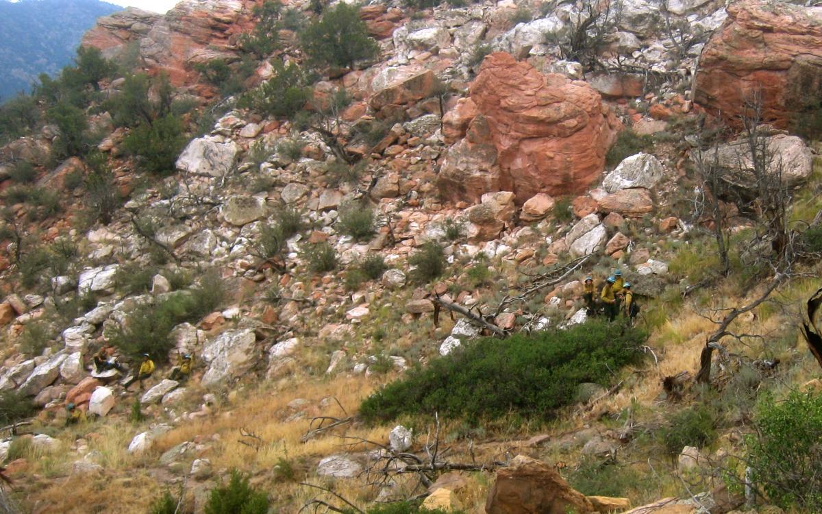 A rocky hillside with large boulders and sparse vegetation, where a group of individuals in yellow and blue climbing gear are navigating the terrain. Oil Well Flats mountain bike trail.