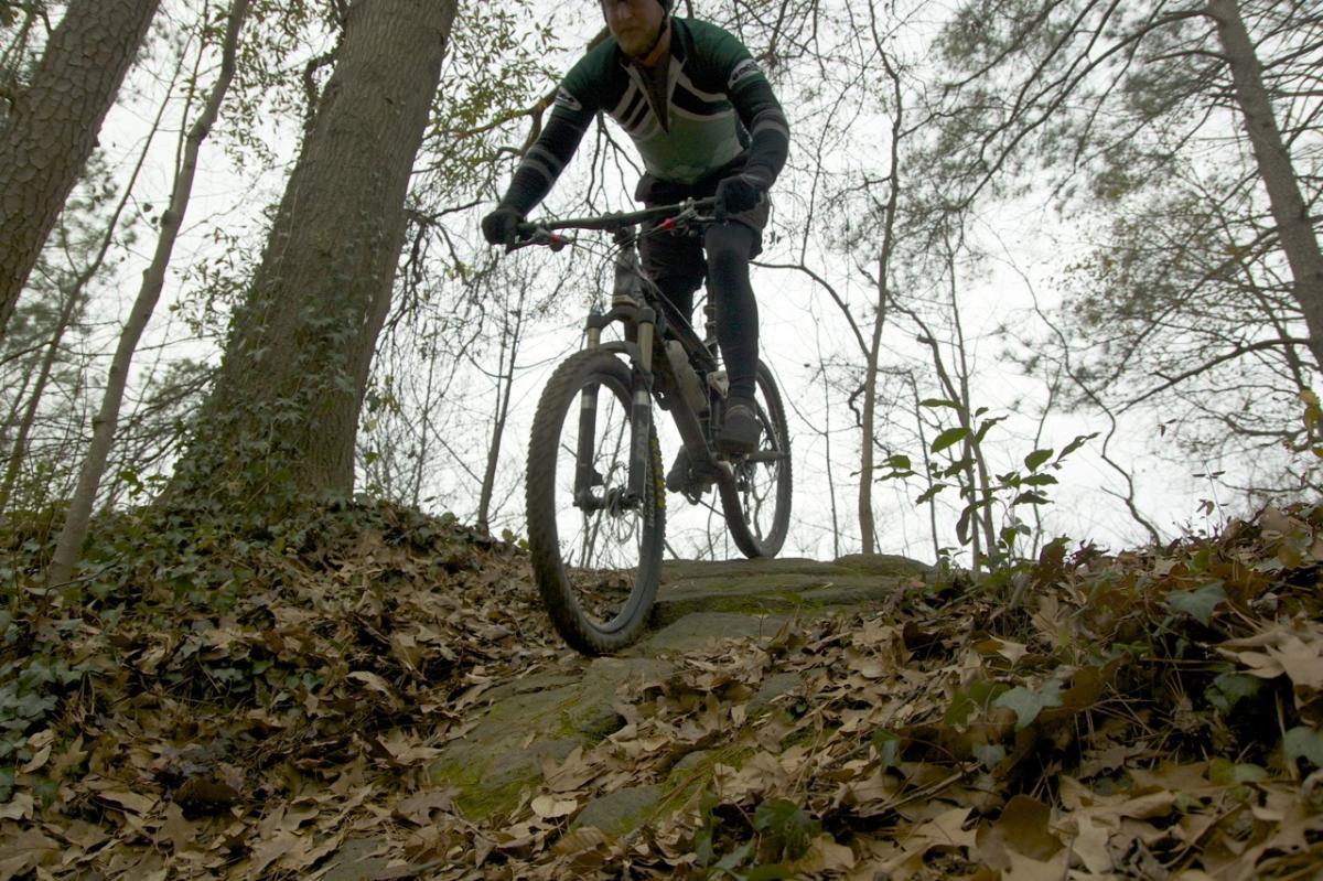 A mountain biker navigating a rocky trail through a wooded area, surrounded by trees and fallen leaves, under a cloudy sky. Ira B Melton mountain bike trail.