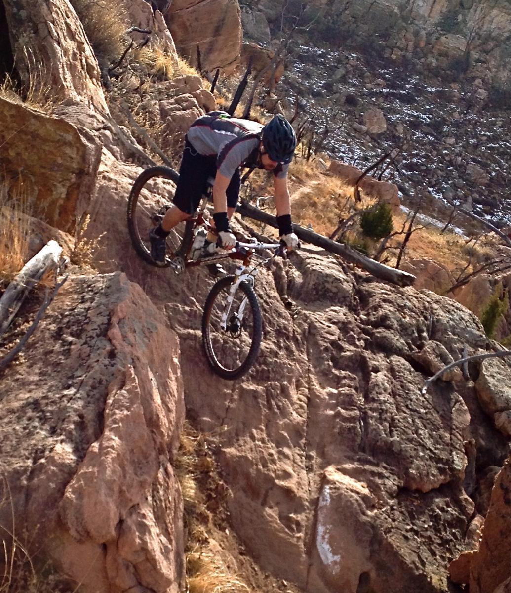 A mountain biker navigates a rocky trail on a steep terrain, wearing a helmet and athletic gear. The surroundings feature rugged rock formations and sparse vegetation, indicating an outdoor adventure in a natural environment. Oil Well Flats mountain bike trail.