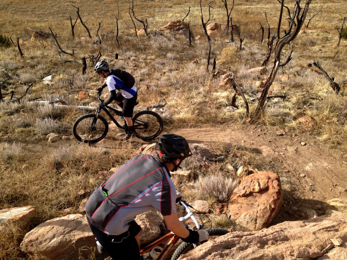Two mountain bikers navigating a rocky trail in a dry, grassy landscape. One cyclist is riding downhill while the other is climbing a rocky section. The surroundings include sparse vegetation and some burnt trees, indicating a rugged outdoor environment suitable for biking. Oil Well Flats mountain bike trail.