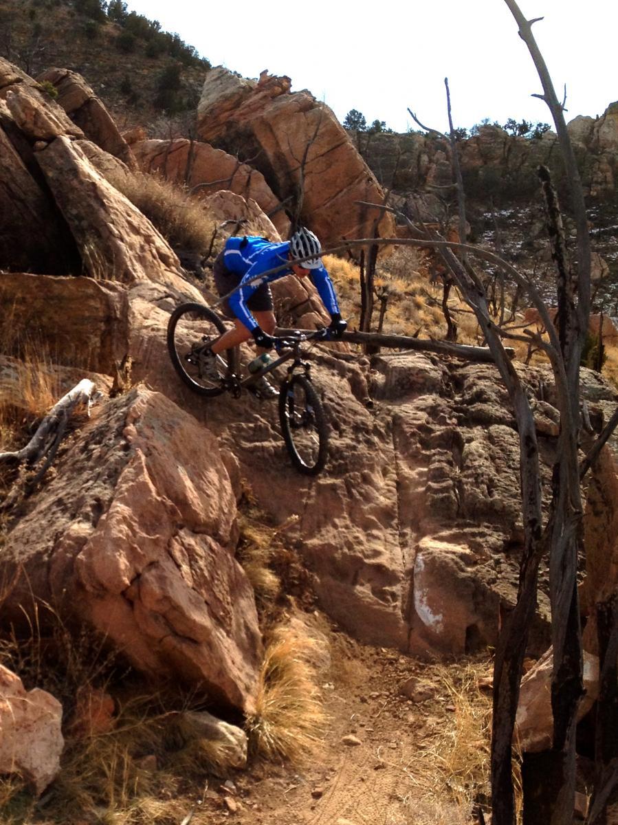 A mountain biker skillfully navigates rocky terrain, leaning forward as they descend a steep, uneven path surrounded by boulders and sparse vegetation. The cyclist is wearing a blue long-sleeve jersey and a helmet. Oil Well Flats mountain bike trail.