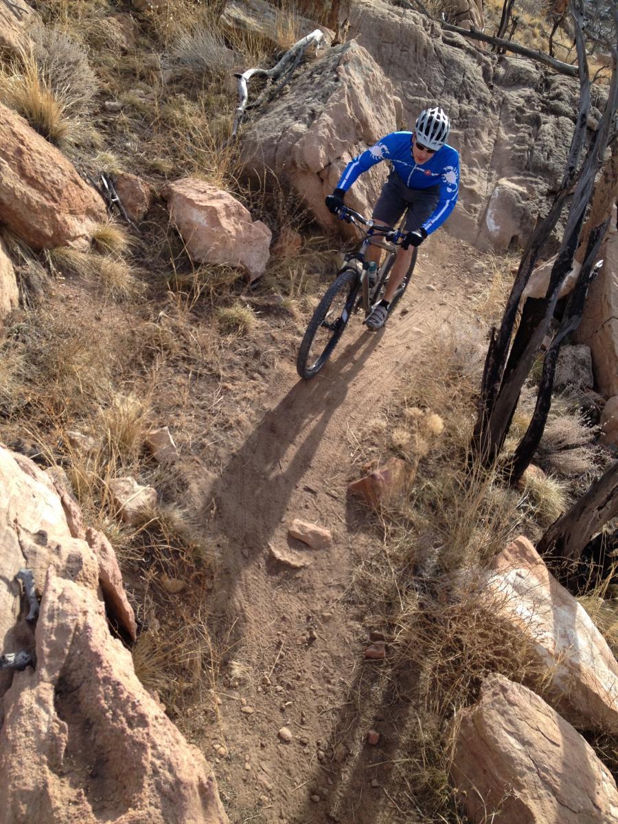 A mountain biker navigates a rocky trail surrounded by dry grass and sparse vegetation, wearing a blue jersey and helmet. The perspective is from above, showcasing the challenging terrain and the biker's focused riding. Oil Well Flats mountain bike trail.