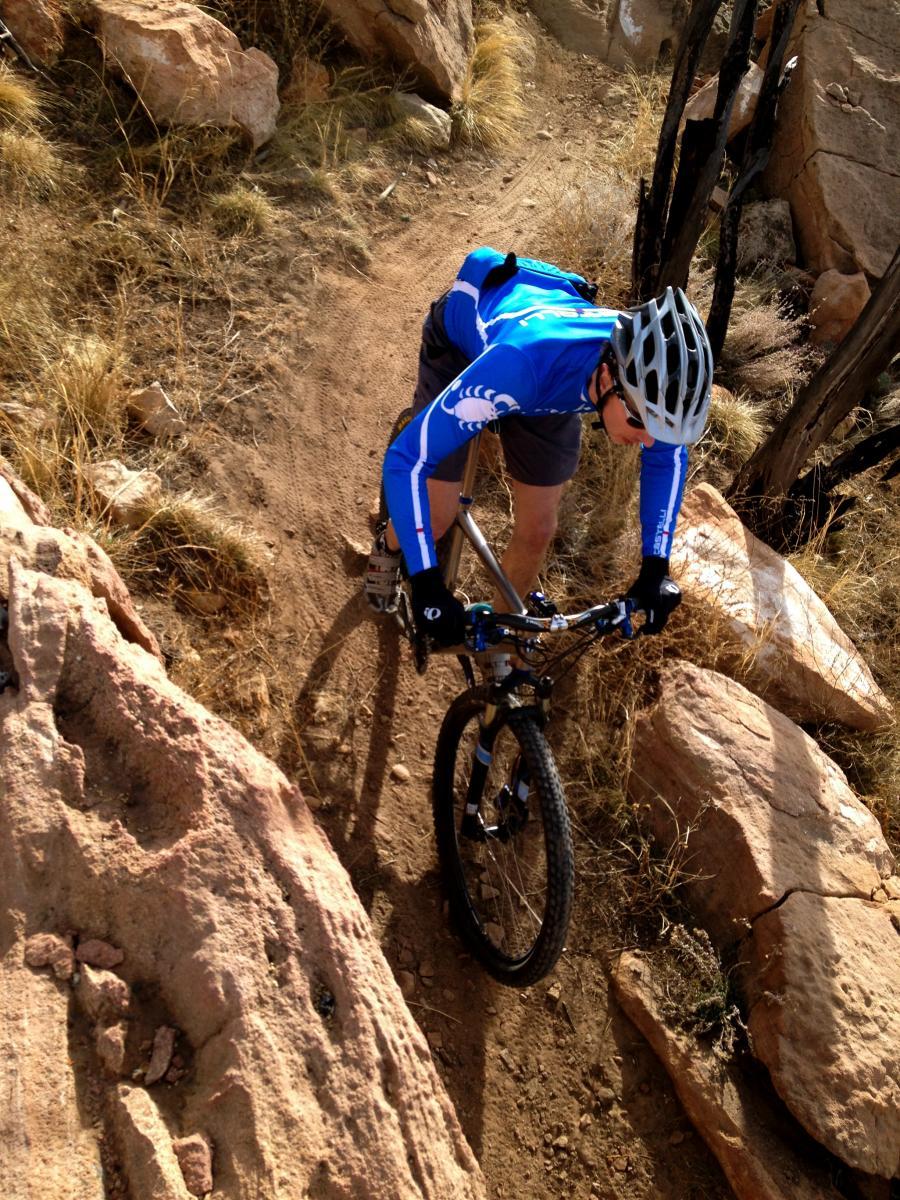 A cyclist in a blue long-sleeve jersey navigates a rocky trail on a mountain bike, surrounded by dry grass and boulders. The rider leans forward, focused on the path ahead. Oil Well Flats mountain bike trail.